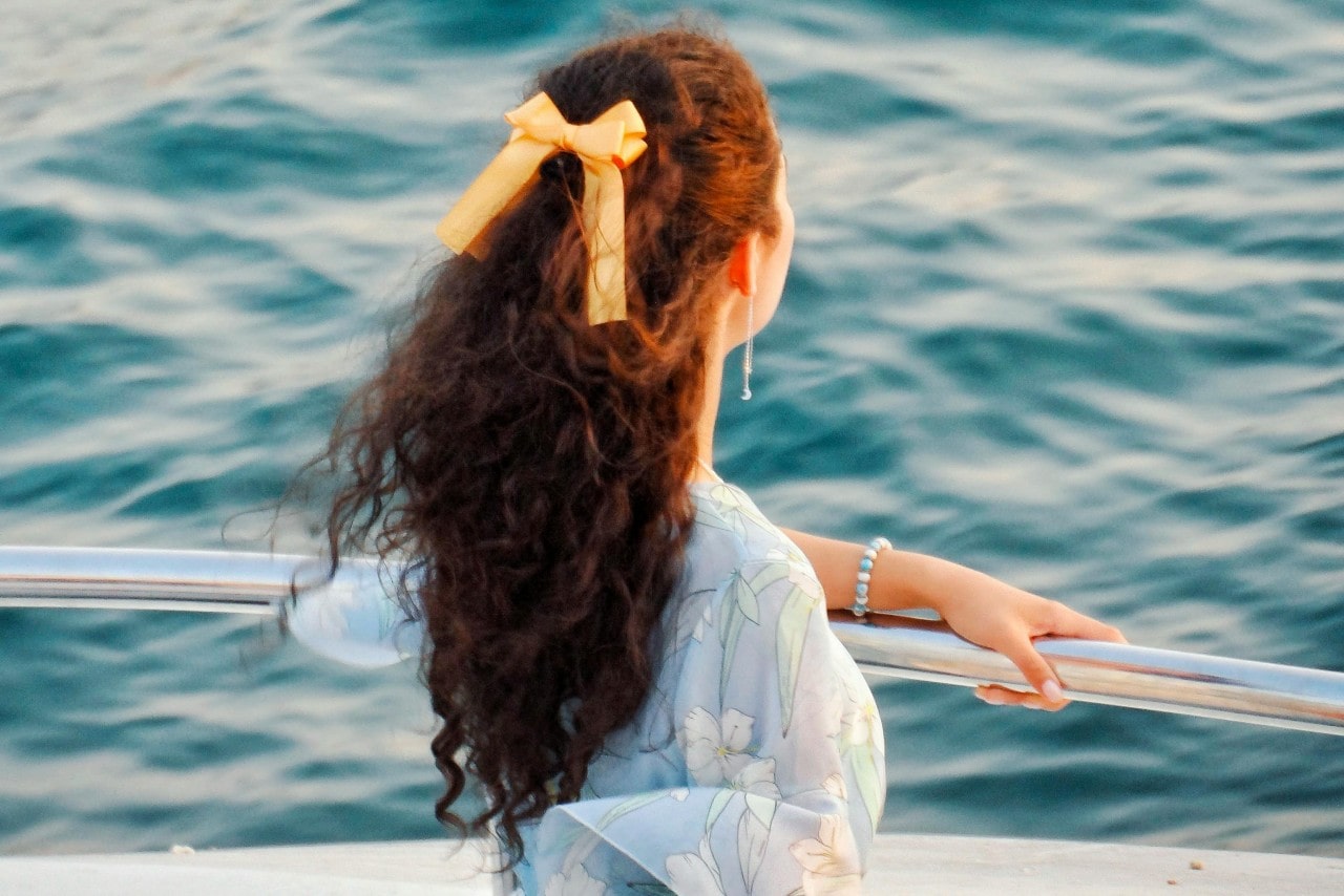 A woman wearing a blue floral dress and an orange ribbon in her curly hair viewing a lake from boat railing.