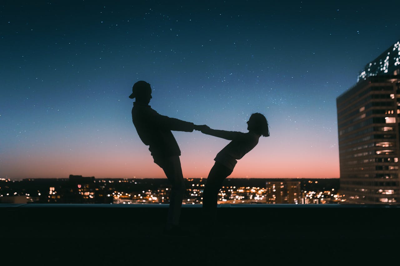 A young couple happily dancing on the rooftop of a hotel among a city skyline as the sun sets.