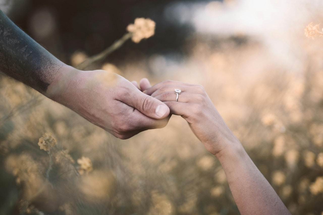 A close-up of a newlywed couple holding hands within a field of golden flowers, with emphasis on her white gold solitaire engagement ring.