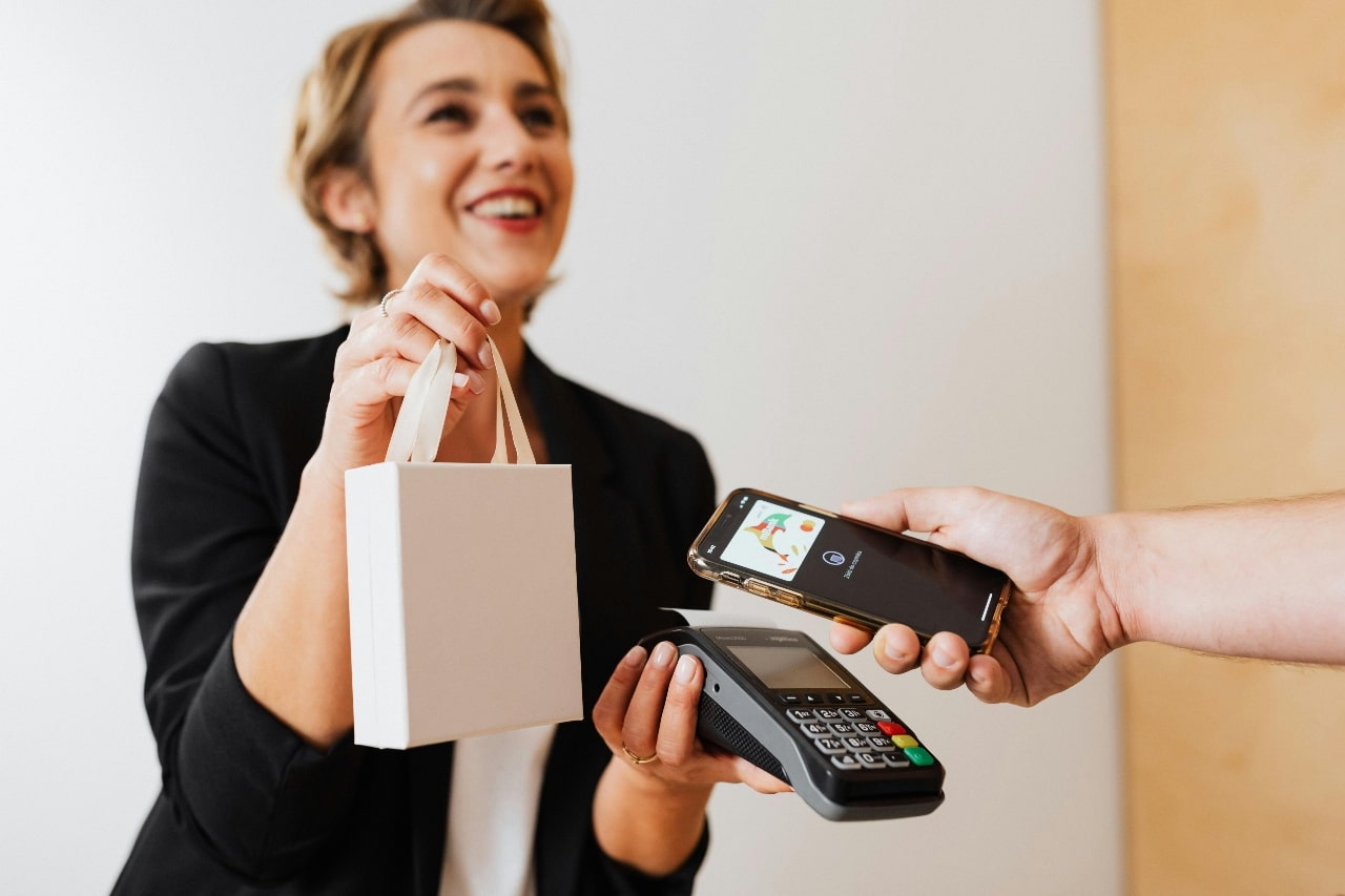 A smiling woman in a blazer hands a small white bag while a person holds a smartphone near a card reader, illustrating contactless payment.