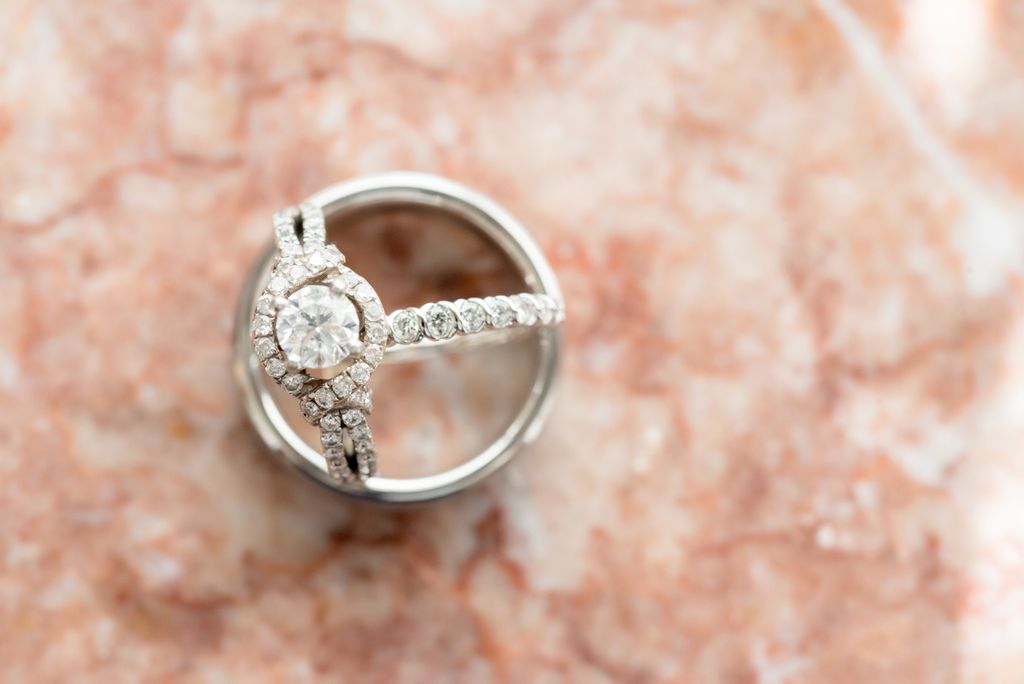 A top-down close-up of an ornate halo engagement ring and matching wedding bands atop a marble surface. A top-down close-up of an ornate halo engagement ring and matching wedding bands atop a marble surface.