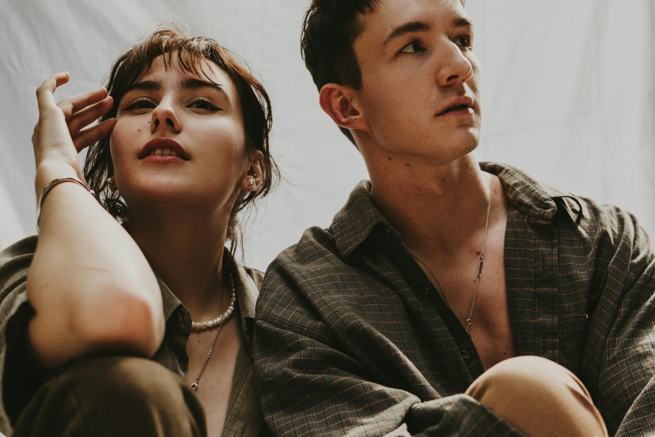A woman adorned with pearl jewelry sits beside a man in a checkered shirt wearing a minimalist chain necklace, both styled in neutral hues.