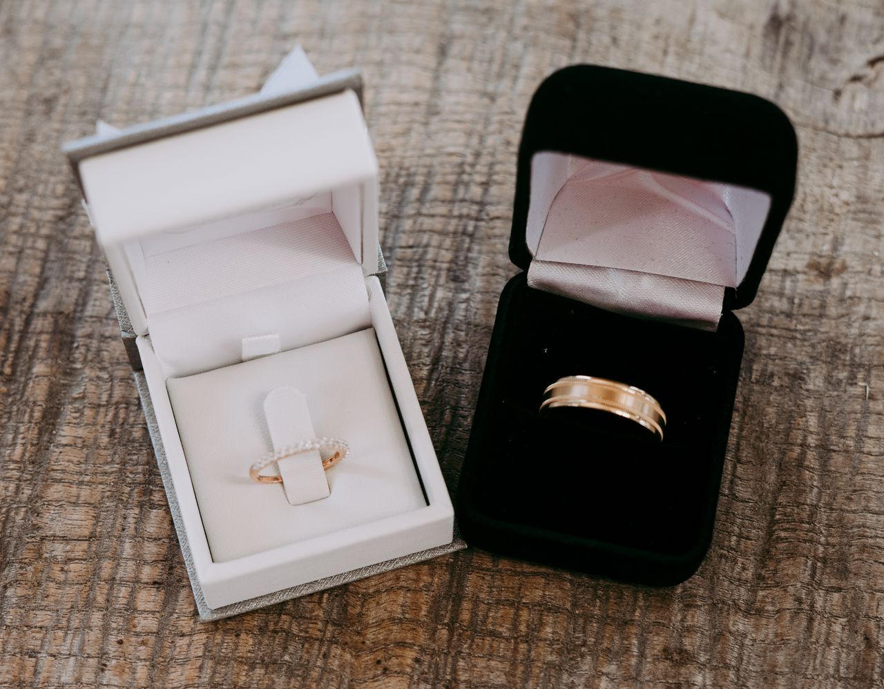 A close up of two velvet jewelry boxes with groom’s and bride’s yellow gold wedding bands, one is white and the other is black, on a wooden surface.