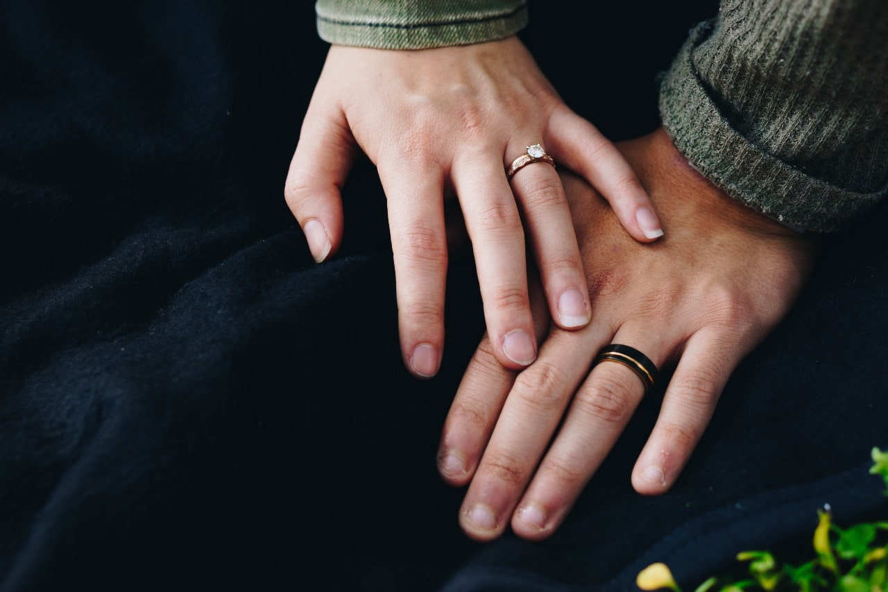 A close up of a couple’s hand on the blue blanket, woman’s hand adorned with rose gold solitaire engagement ring, while man’s with mixed metal wedding band.