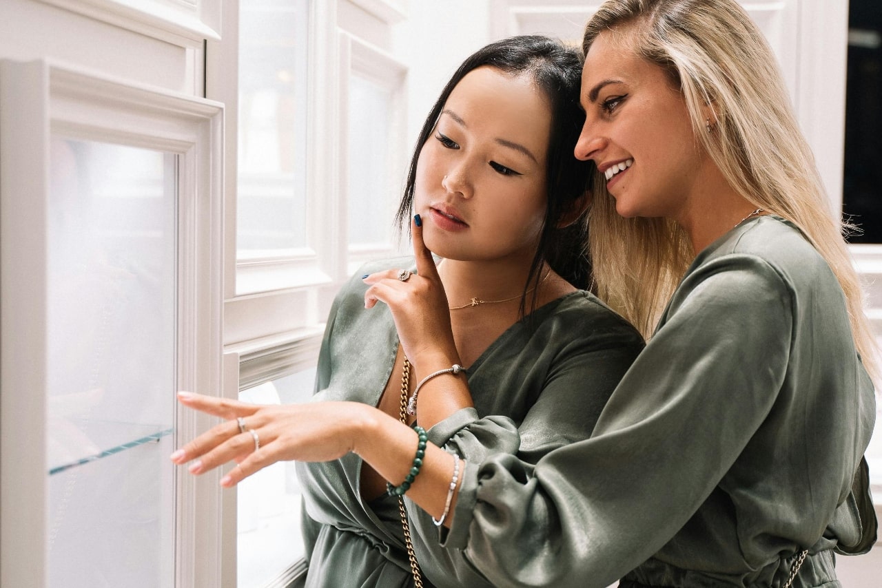 Two women in olive satin blouses admire jewelry in a store display.