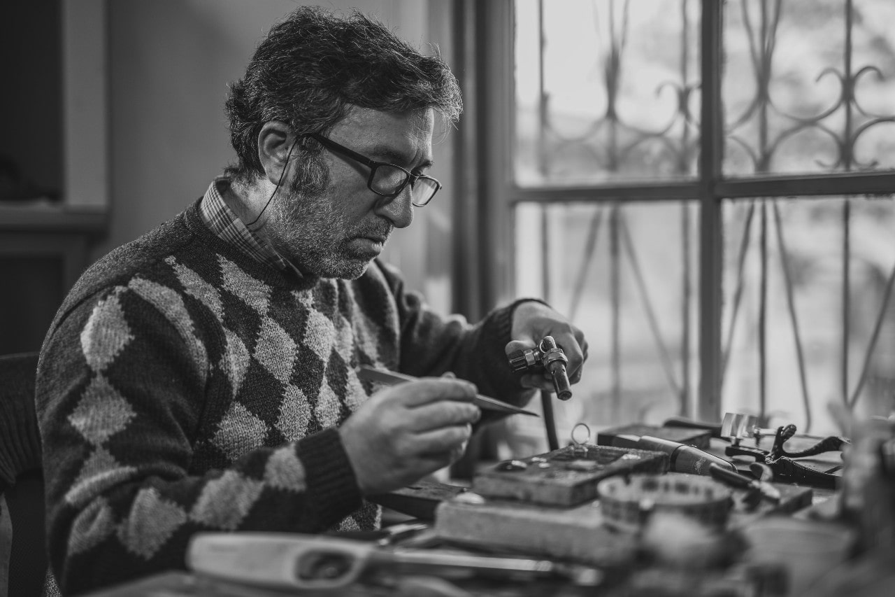 A focused artisan with glasses works on a detailed project at a cluttered workbench by a window. The black-and-white tone adds a nostalgic feel.