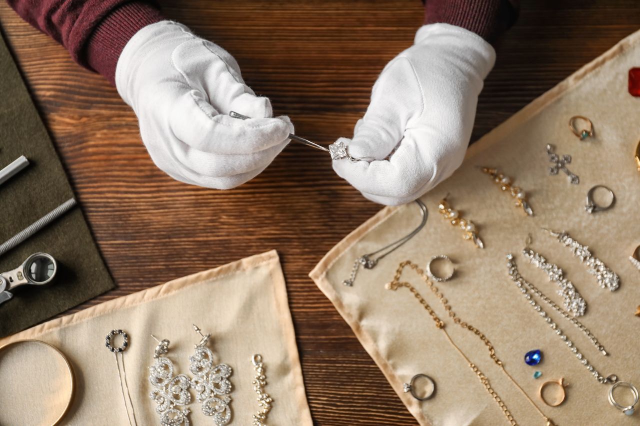 A jeweler wearing white gloves inspecting a diamond ring with tweezers, with a multitude of fine pieces before them placed on protective cloths.