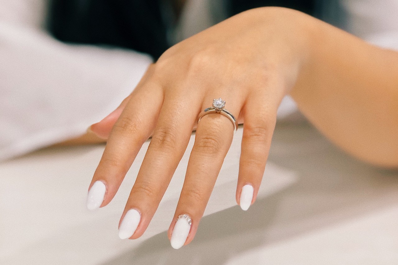 A hand with elegant, white manicured nails displays a diamond engagement ring, set against a soft, blurred background.